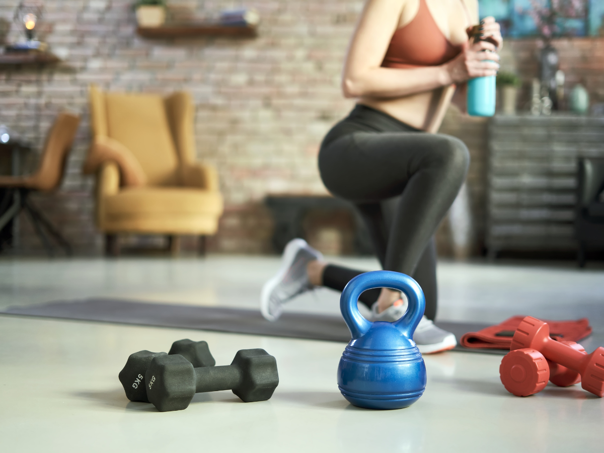 The image of a spacious living room with a brick wall in the background a mustard cushion chair and a woman in black leggings working out on a yoga mat, drinking from an aquamarine bottle, and different kinds of weights on the floor.