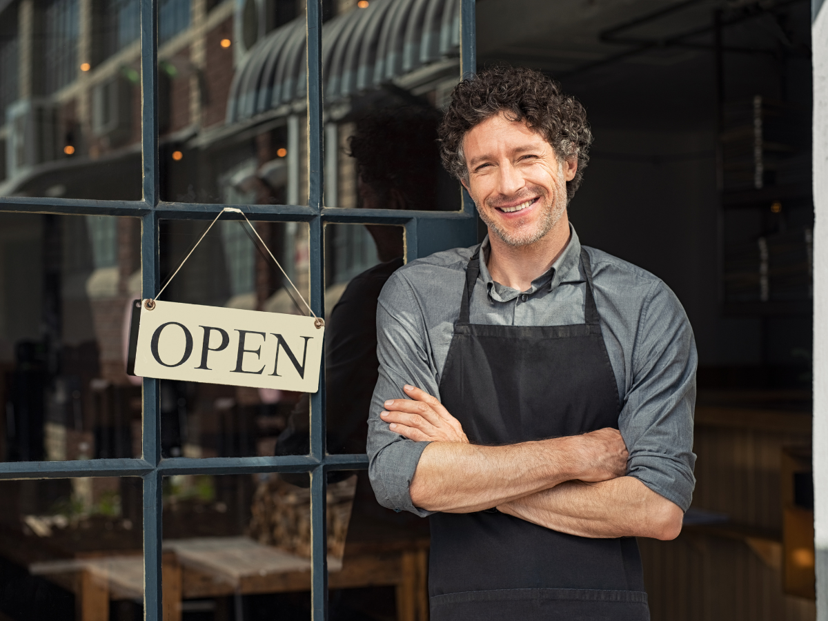 A smiling middle-aged man with a black apron, laying down a restaurant glass door with an open sign.