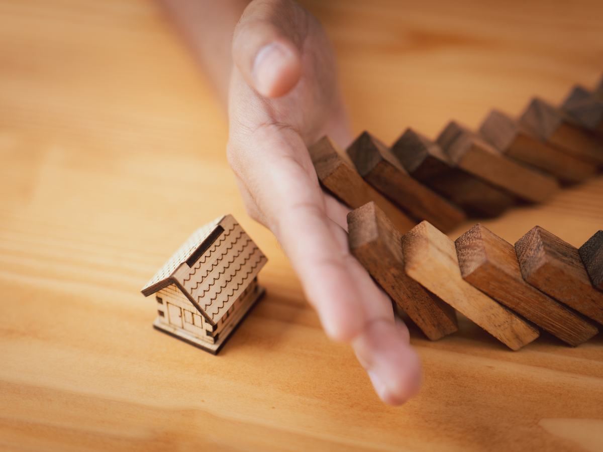 A hand stopping a dominoes falling in a row over a home doodle on top of a light-colored wood table.