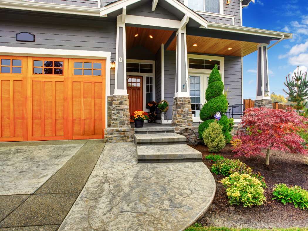 Photo of the curb appeal of a house with a well-maintained driveway, some bushes, and cypress pines around the entrance, and a small front porch with colorful flower pots before the front door.