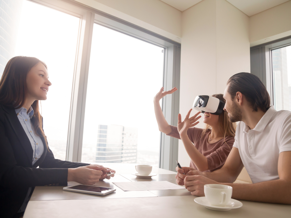 A real estate agent talking with a couple in a building office, they're around a table with coffee and documents, and the woman has 3D virtual reality glasses on, to take the virtual tour of a home for sale.